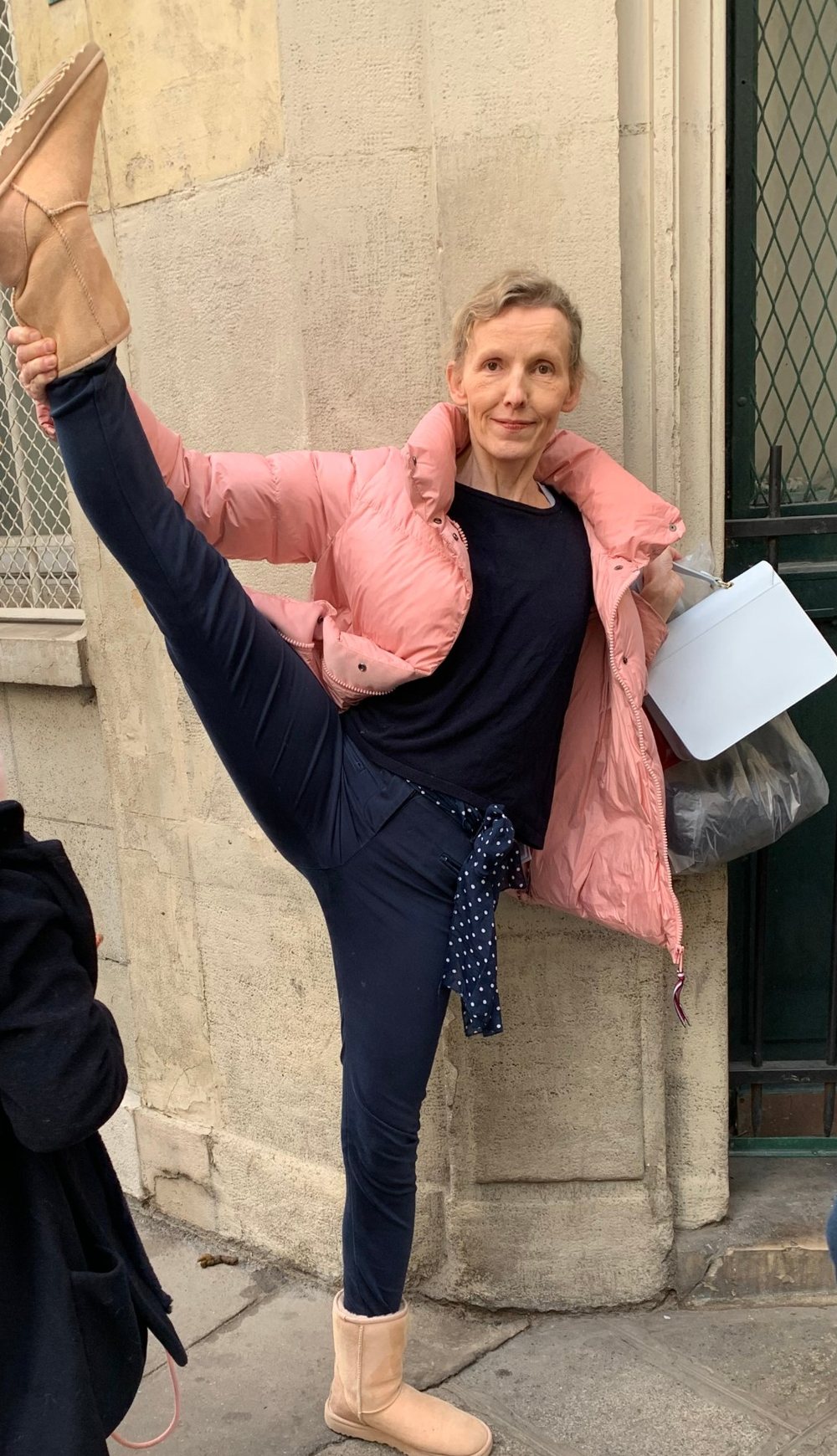 Ariane Haneveld in rosa Daunenjacke hält ihr Bein im Stehen senkrecht nach oben, an einer Hauswand in Paris